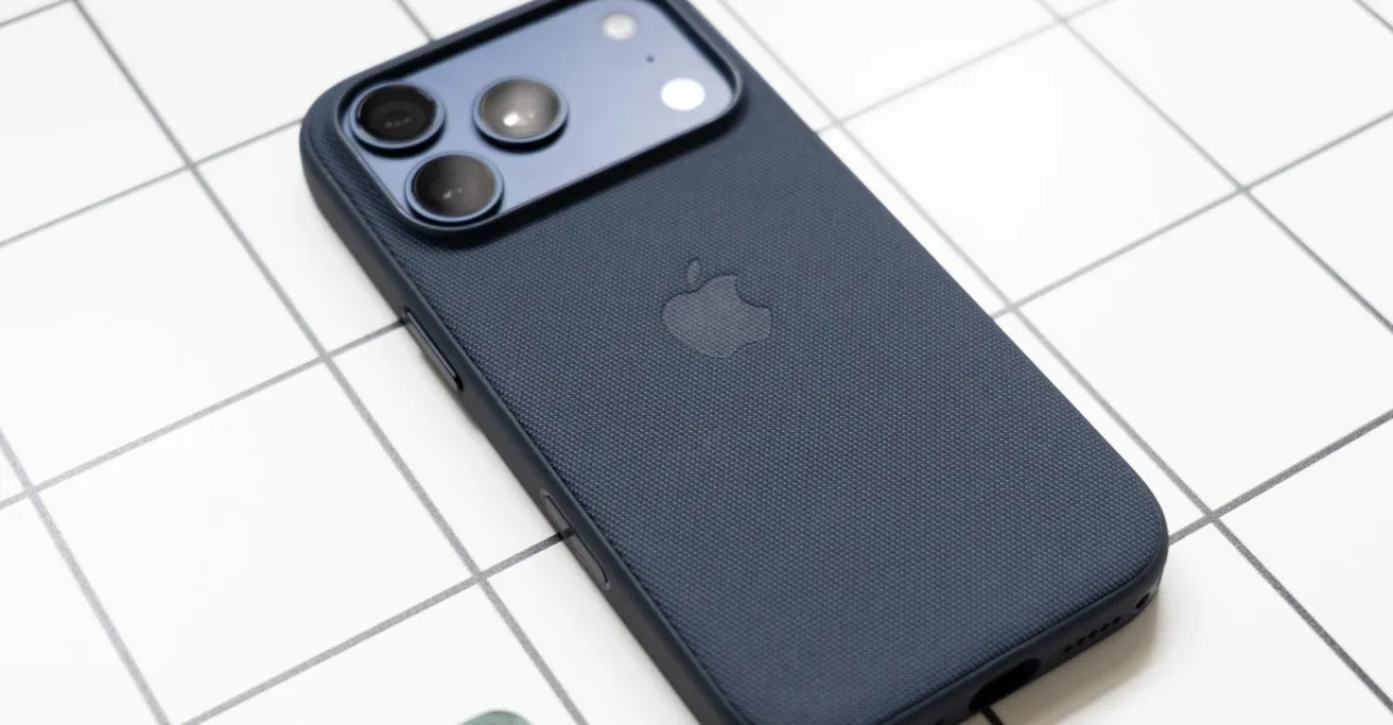 Um homem usando iPhone enquanto faz agachamento com halteres, ao fundo um MacBook aberto