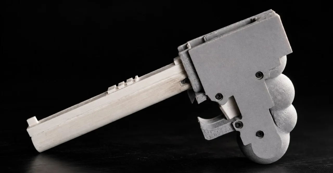 Cody Wilson exibe a pistola WASP, um rifle compacto com acabamento metálico e tela de código ao fundo