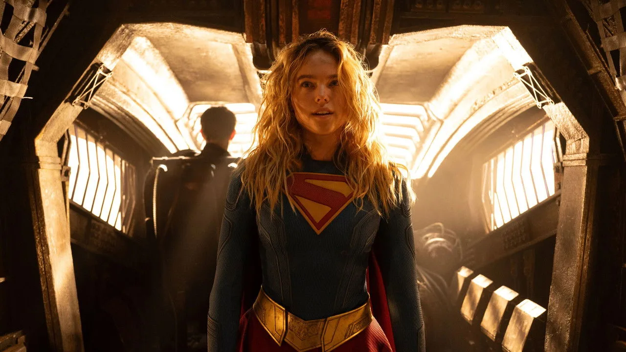 Kara Zor‑El em traje azul, levantando halteres numa academia, com capa ao fundo e pôster do Superman na parede