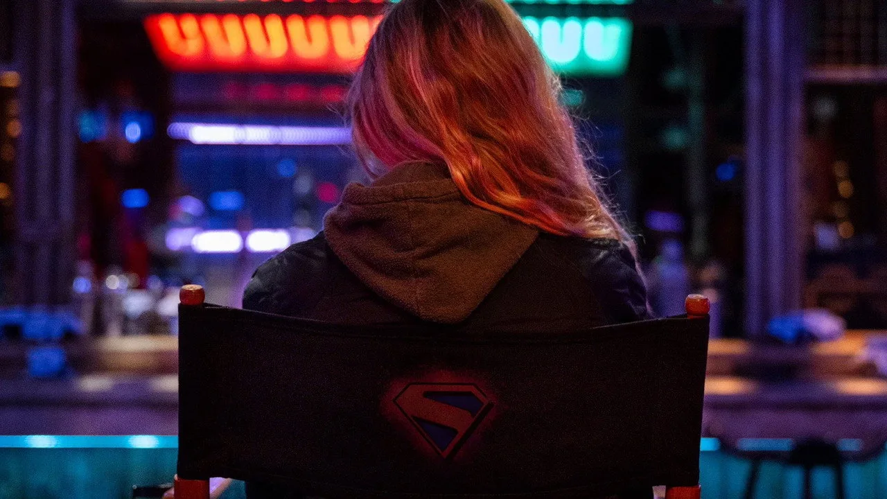 Mulher musculosa vestindo camiseta da Supergirl faz agachamento com barra e halteres ao fundo