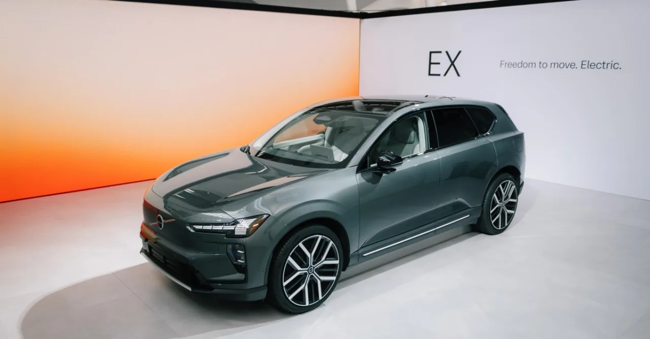 Motorista em postura ergonômica, usando smartwatch, ao lado de carro Volvo com tela exibindo alerta de pedestre
