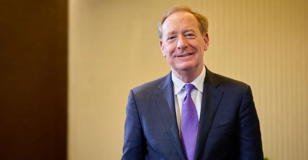 Brad Smith, em terno, aparece ao lado do logotipo da Microsoft em um fundo corporativo