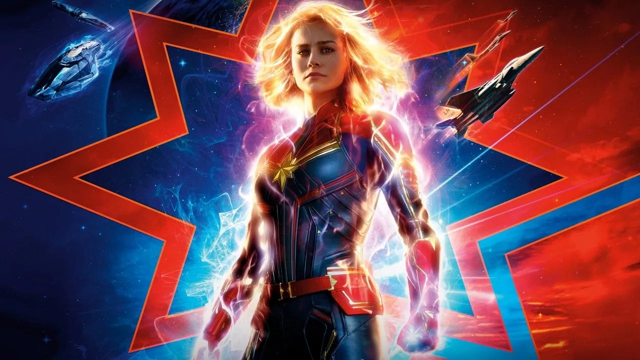 Mulher musculosa vestindo o uniforme da Capitã Marvel levanta halteres vermelhos ao lado de um rack de pesos