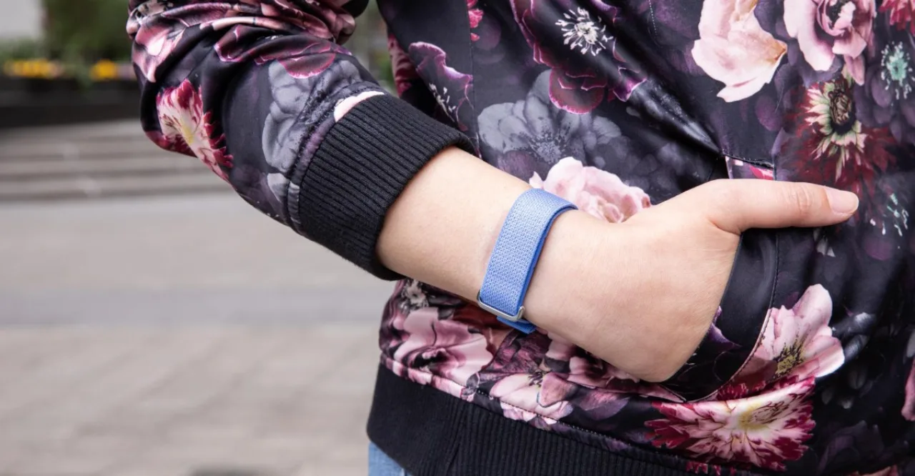 Pessoa em roupa esportiva, usando Fitbit Air no pulso, olhando tela do celular com gráficos de sono e batimentos, ao fundo praia ensolarada