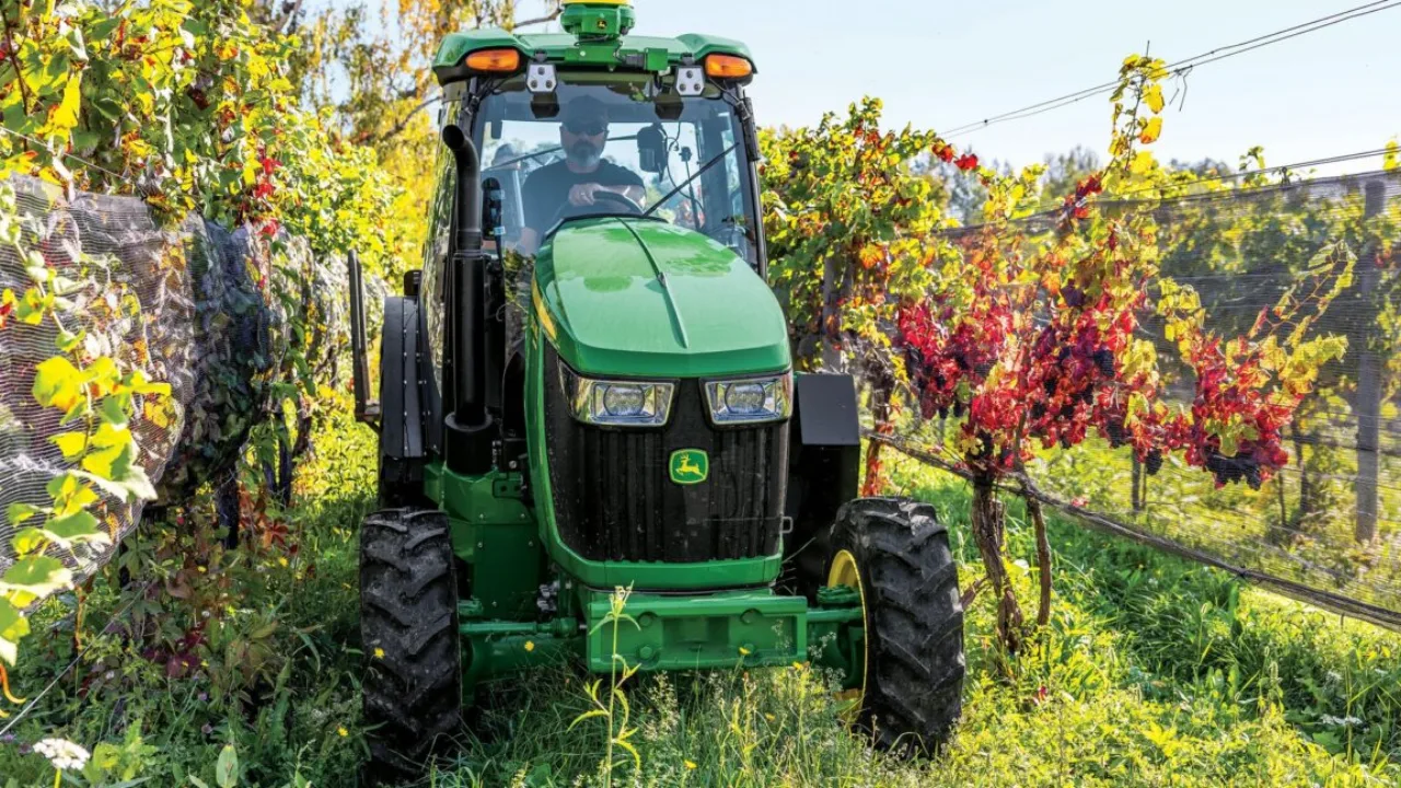 Trator John Deere 5130ML verde‑amarelo no campo, técnico ao lado com tablet e logo da marca ao fundo