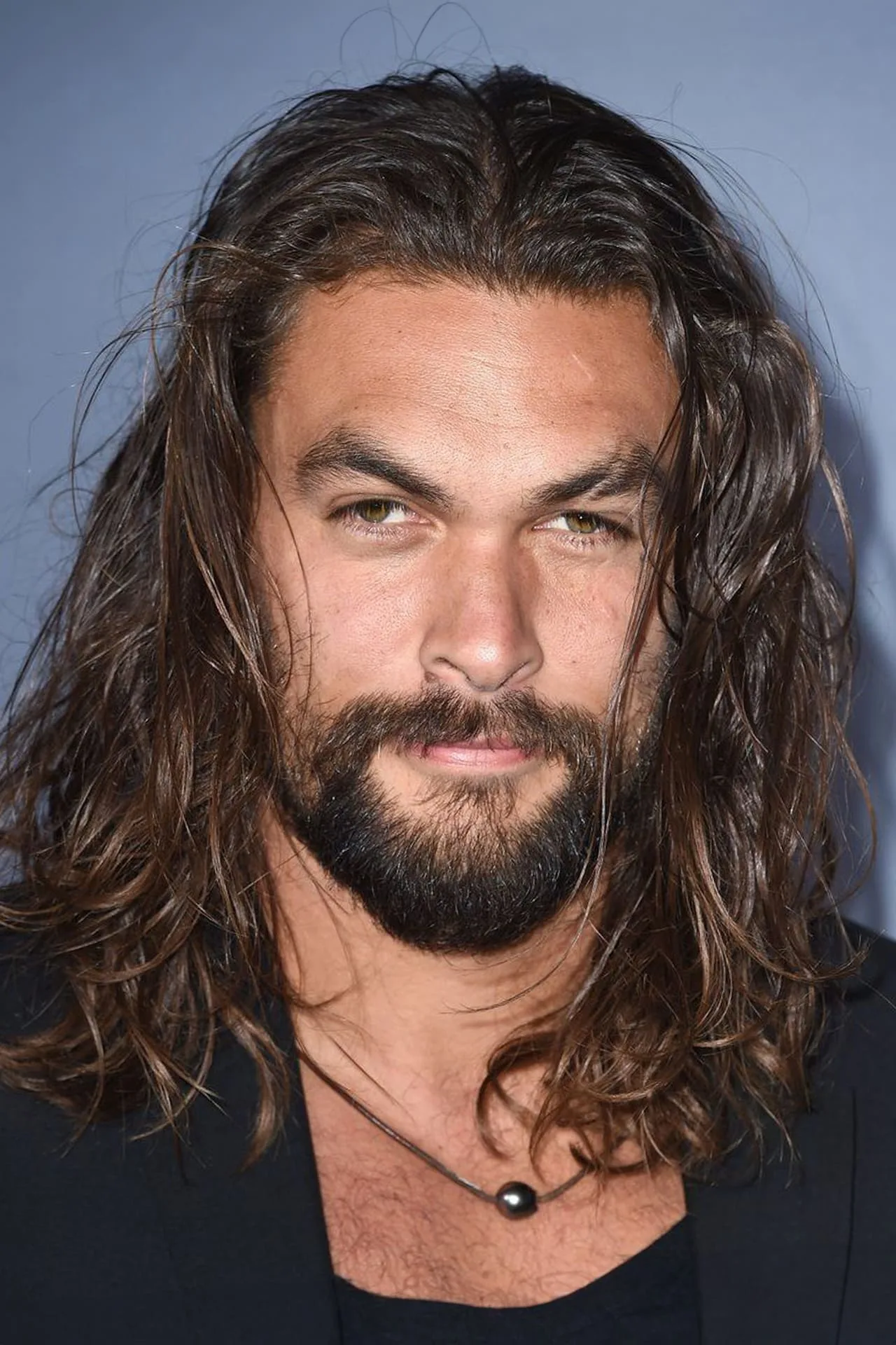 Jason Momoa malhando com barra e halteres, usando camiseta de super‑herói, suado e focado