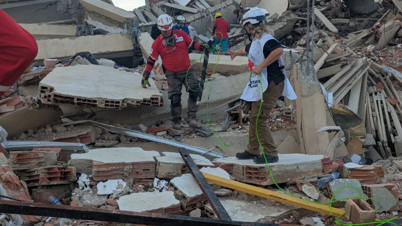 Robô serpente cinza deslizando entre escombros de concreto, com capacete de resgate ao fundo
