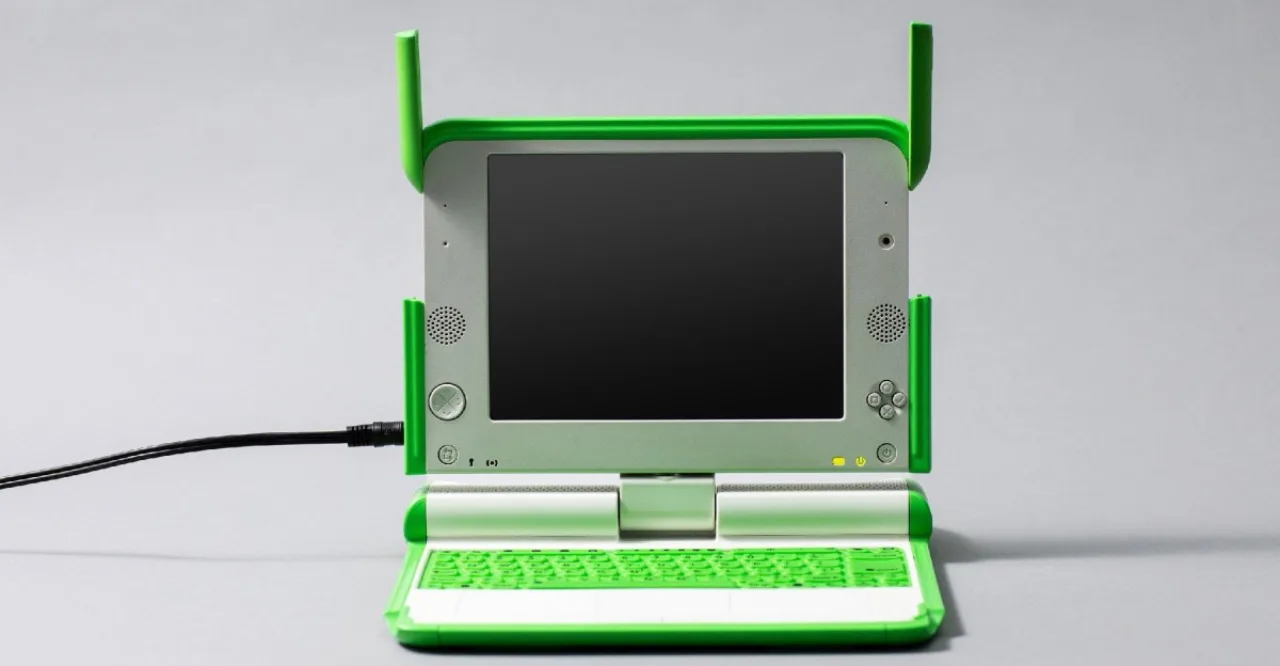 Criança segurando um laptop amarelo da OLPC com o logotipo ao fundo de uma sala de aula simples