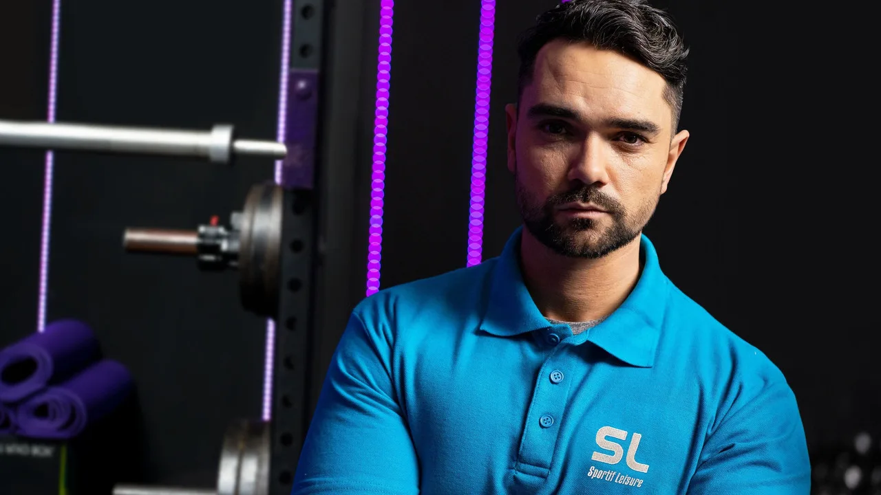 Mulher em roupa esportiva faz agachamento com kettlebell num parque, segurando garrafa d’água ao lado