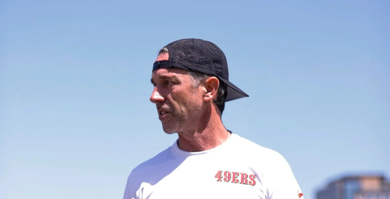 Kyle Shanahan vestindo roupa de treino, segurando um volante de Tesla com o logo do Autopilot ao fundo