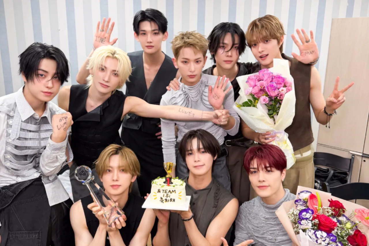 &TEAM celebra no palco de Music Bank segurando o troféu, rodeado por luzes neon e o logo do programa ao fundo