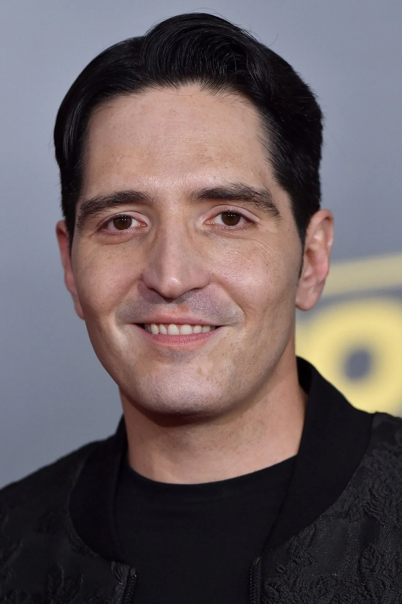 David Dastmalchian vestindo roupa de treino, segurando halteres ao lado de um quebra‑cabeça de peças de Charada