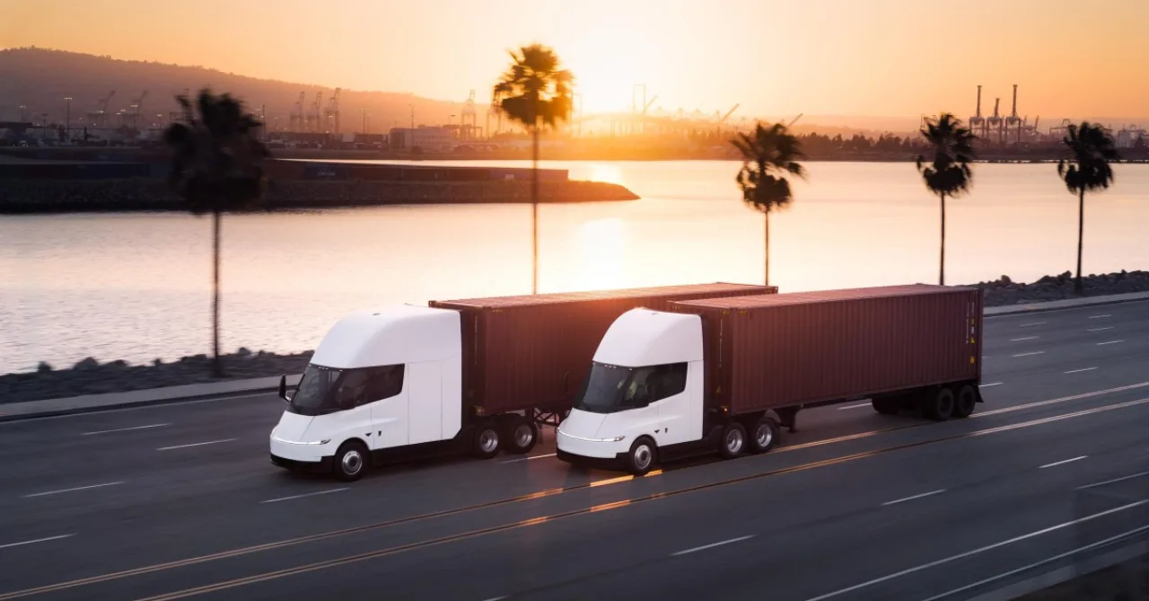 Tesla Semi azul estacionado ao lado de um robô de montagem, com o logotipo da Tesla ao fundo na fábrica