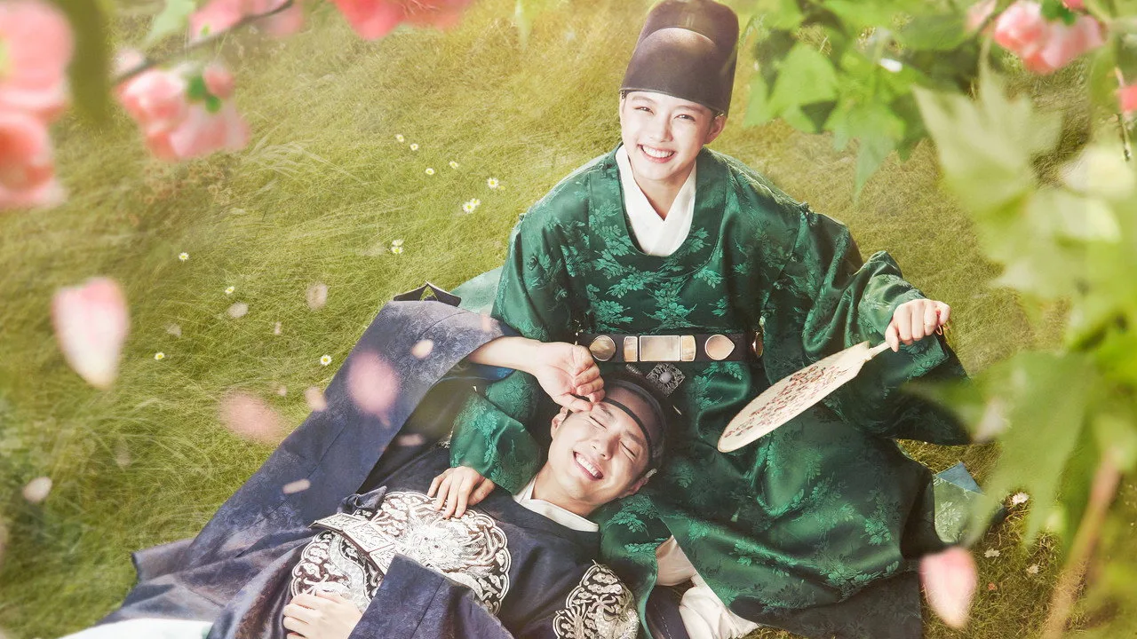 Jovem em roupa esportiva faz alongamento no sofá, TV mostra cena de Love In The Moonlight e chá verde ao lado