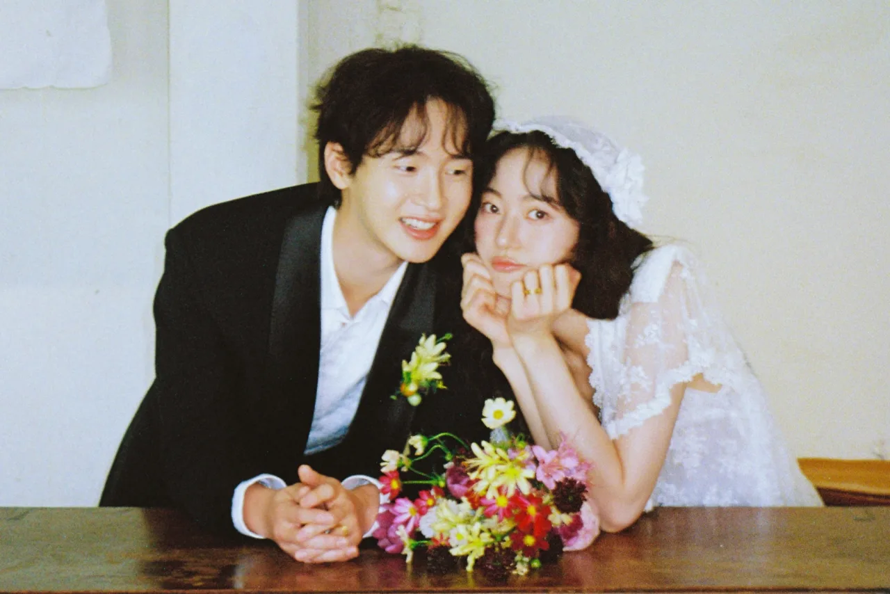 Jang Dong Yoon e Kim Seung Yoon aparecem de terno e vestido branco, sorrindo em um elegante cenário de casamento