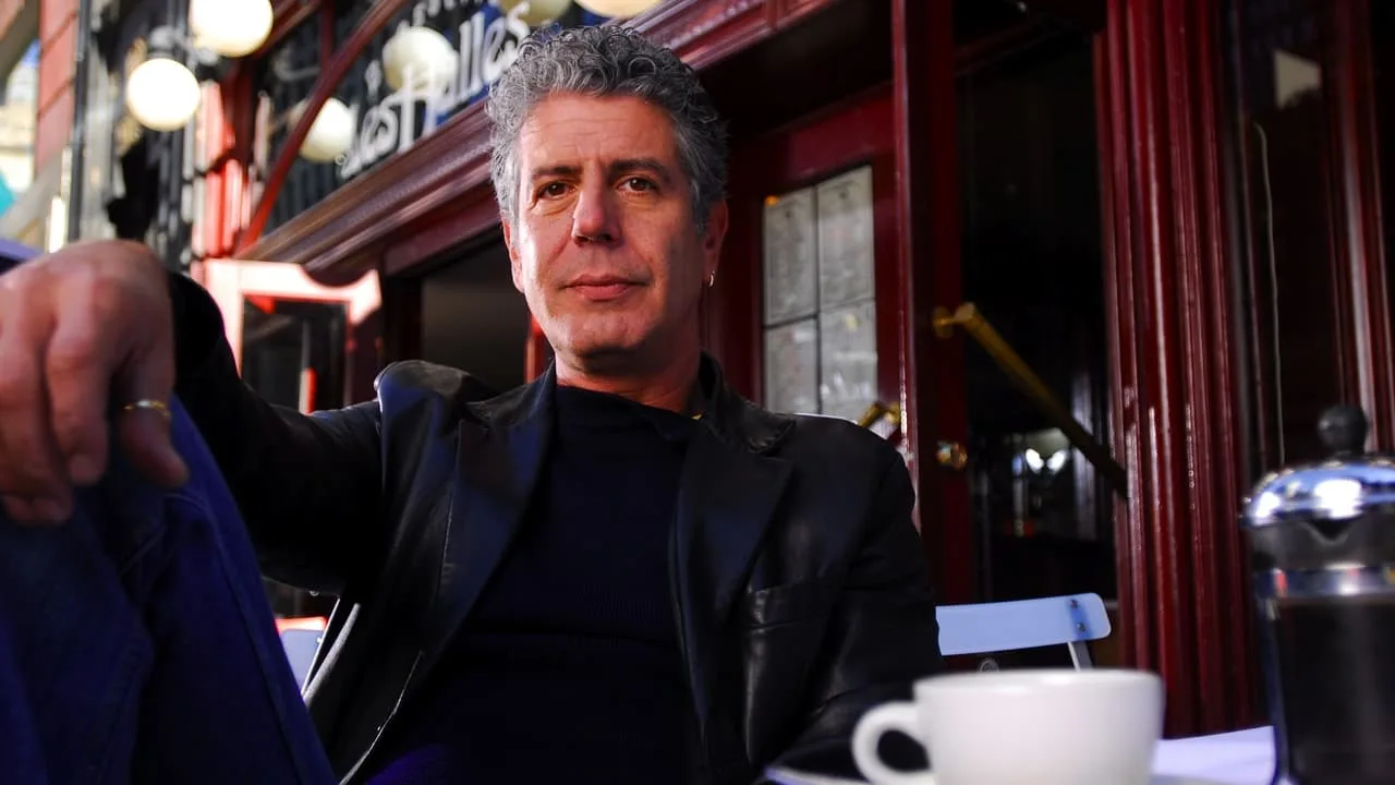 Bourdain segurando uma faca de chef ao lado de um controle de videogame, com fundo de selva tropical