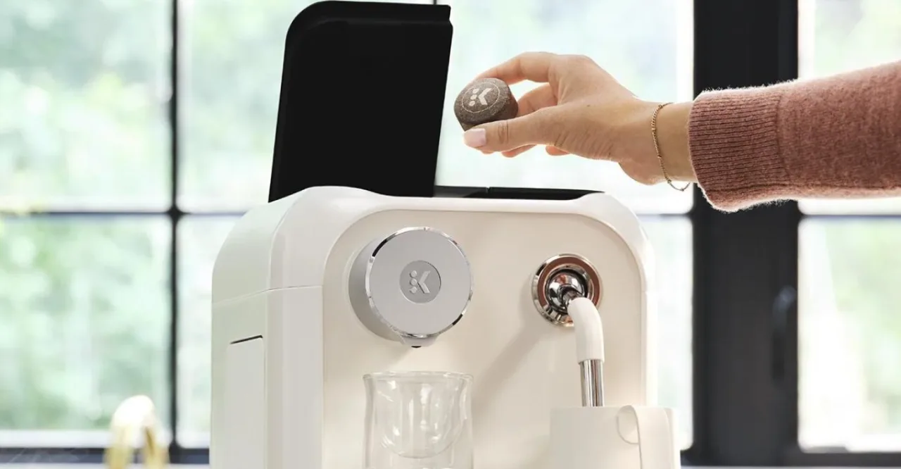 Máquina Keurig Alta ao lado de pucks compostáveis verdes de algas, com logo da marca ao fundo