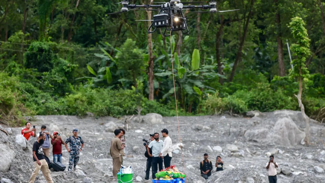 Drone DJI FlyCart sobrevoando montanhas nepalenses, carregando caixa de suprimentos enquanto equipes militares observam