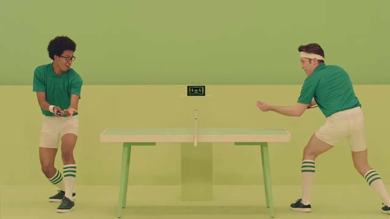 Pessoa jogando videogame em console portátil com controle ergonômico, garrafa de água e frutas sobre uma mesa