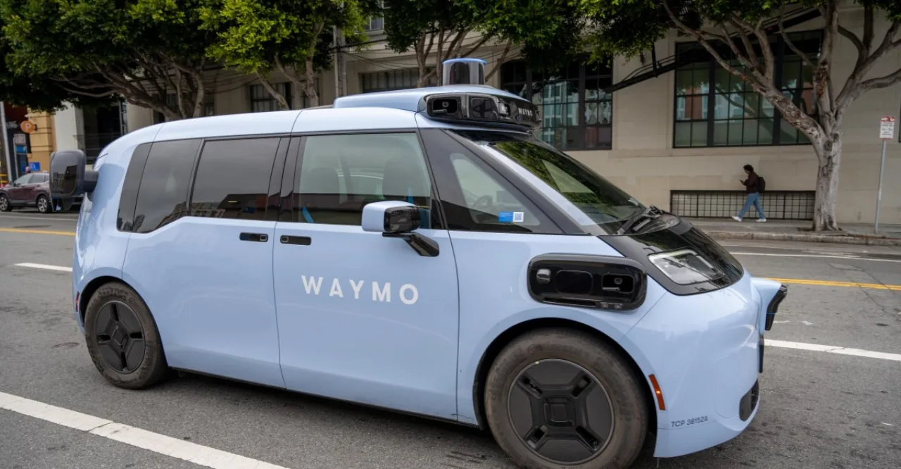 Veículo Waymo Ojai autônomo circula em rua urbana, com logo Waymo visível e edifícios ao fundo