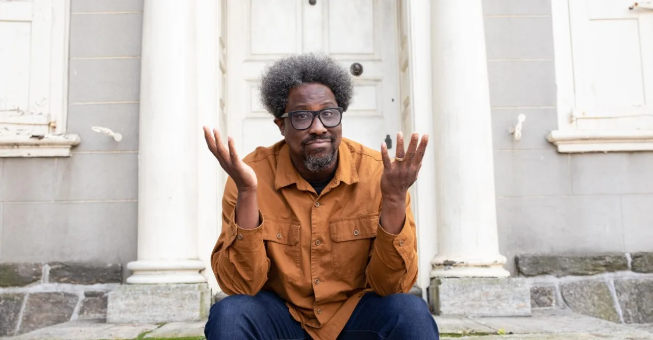 W. Kamau Bell levantando halteres, camiseta com slogan de justiça, ao lado de garrafa d’água e tapete de yoga