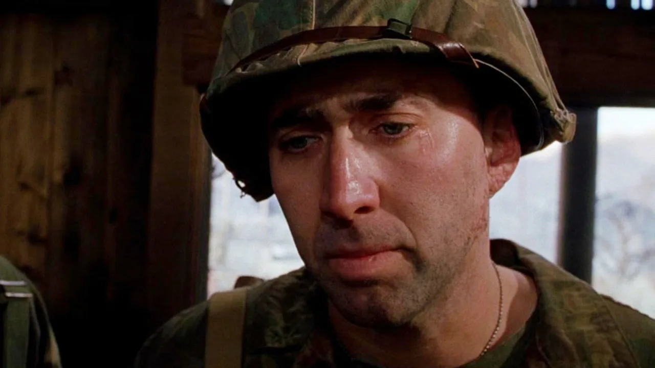 Nicolas Cage suado com fuzil e rádio na selva, destacando o vigor físico e a resistência muscular em cena de combate