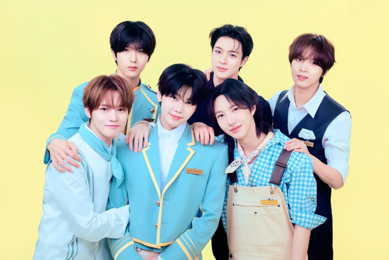 Sete integrantes do NCT DREAM fazendo alongamento em estúdio com colchonetes coloridos e garrafas de água
