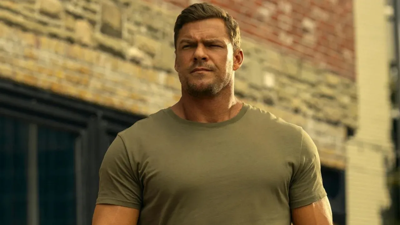Alan Ritchson vestindo roupa de treino, segurando halteres e exibindo músculos ao ar livre
