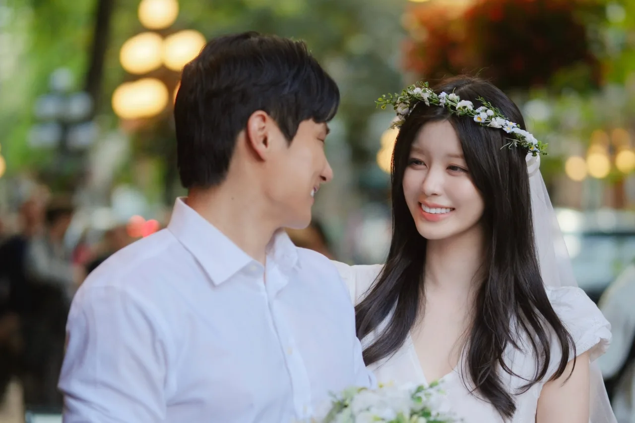 Soyee sorrindo ao lado do noivo, vestindo traje nupcial branco, cercada por flores de lótus e um bolo de frutas frescas