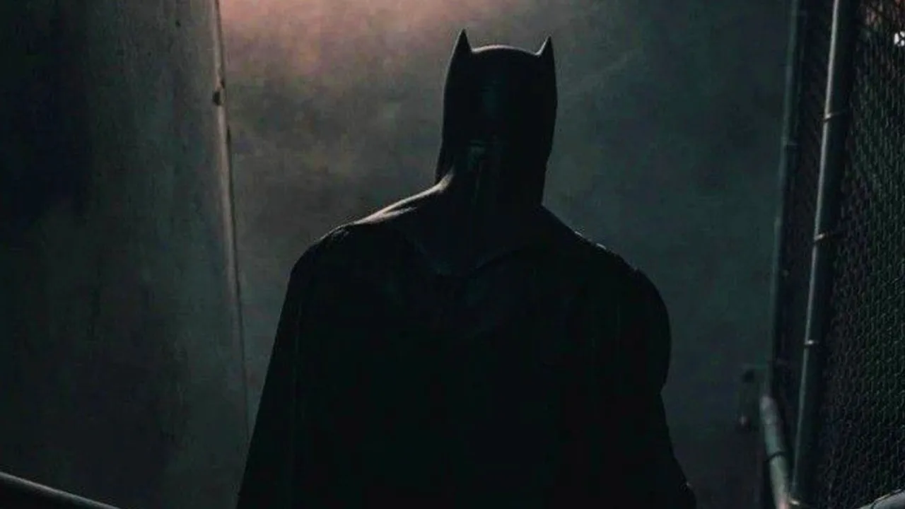 Homem musculoso vestindo capa e máscara do Batman levanta halteres em academia iluminada