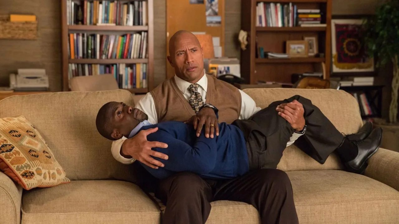 Dwayne Johnson e Kevin Hart levantando halteres lado a lado, sorrindo, em academia iluminada