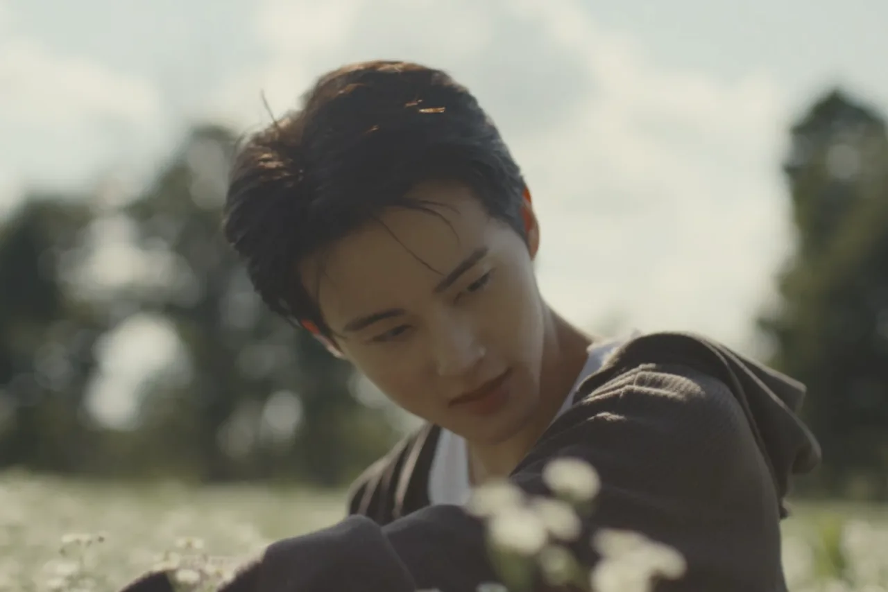 Jovem com fones, fazendo agachamento com halteres, garrafa d'água e laptop exibindo o teaser de Mark Lee