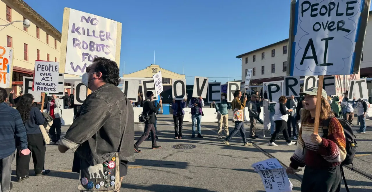 Manifestantes segurando placas com o texto 'People Over Profit' ao redor do palco com o logotipo da OpenAI