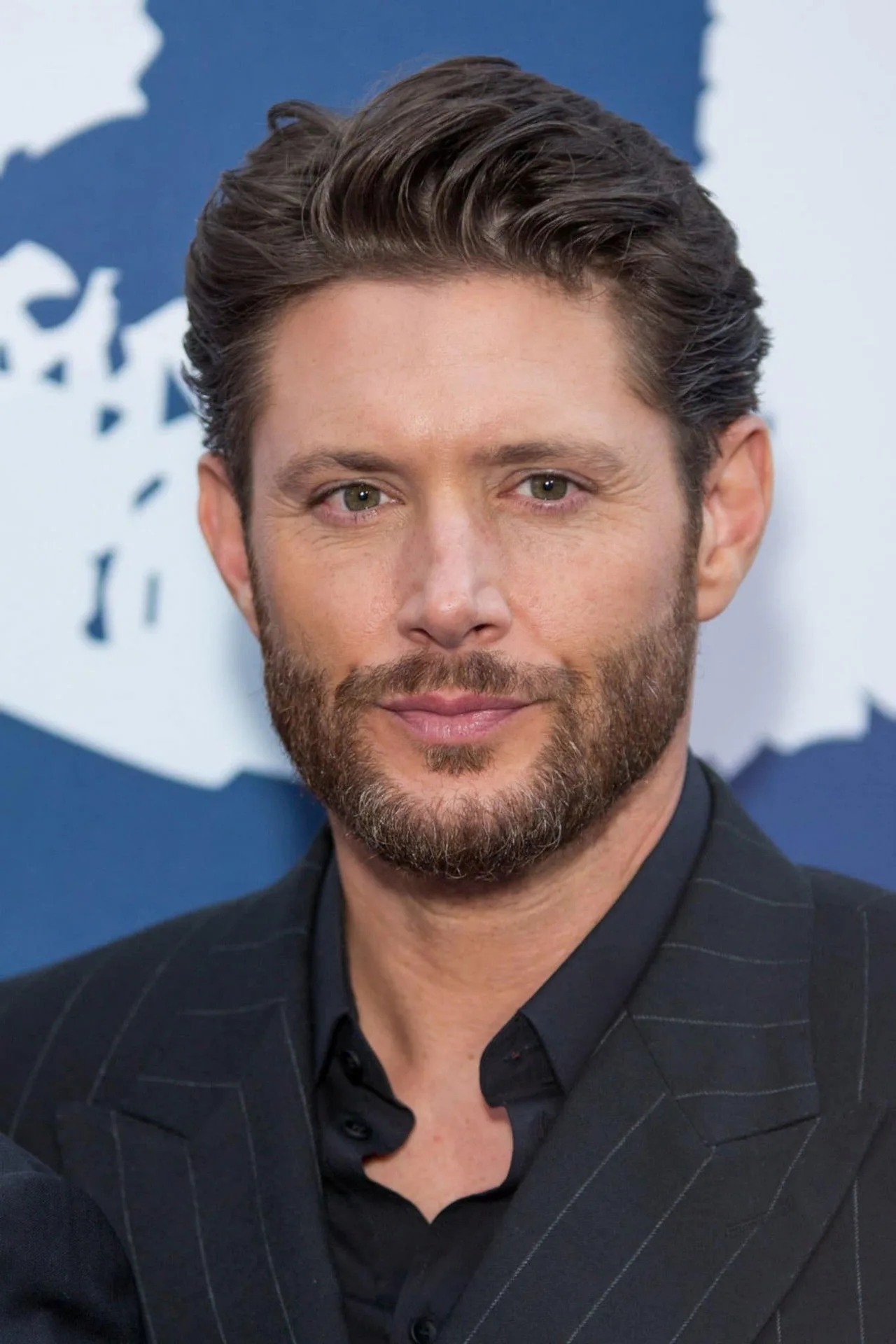 Jensen Ackles em pose atlética e séria, vestindo jaqueta de couro e segurando um equipamento de rastreamento tático