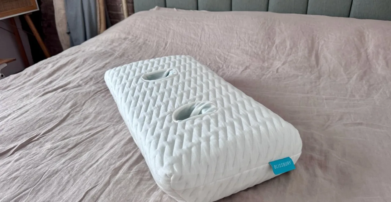 Travesseiro de espuma viscoelástica ao lado de fones Sleepbuds, ambos sobre uma cama com luz suave ao amanhecer