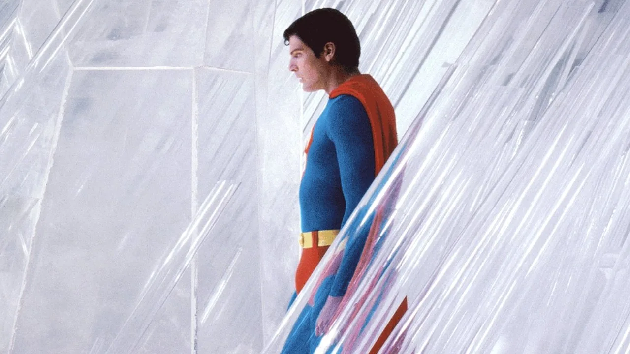 Homem executa o exercício Superman na areia, ao lado de uma prancha de yoga e uma mochila de sobrevivência