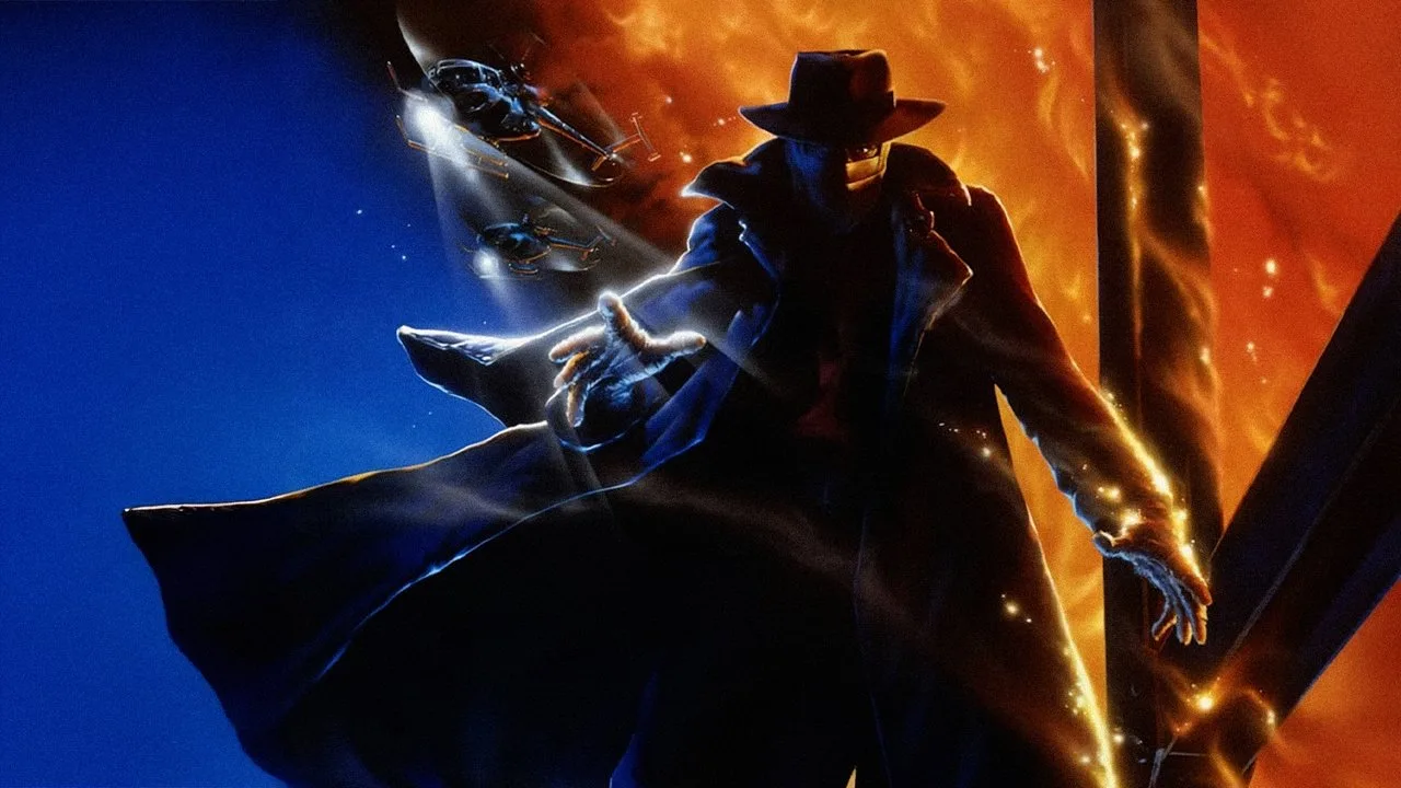 Homem musculoso vestindo capa preta, segurando halteres, com máscara de Darkman ao fundo de laboratório