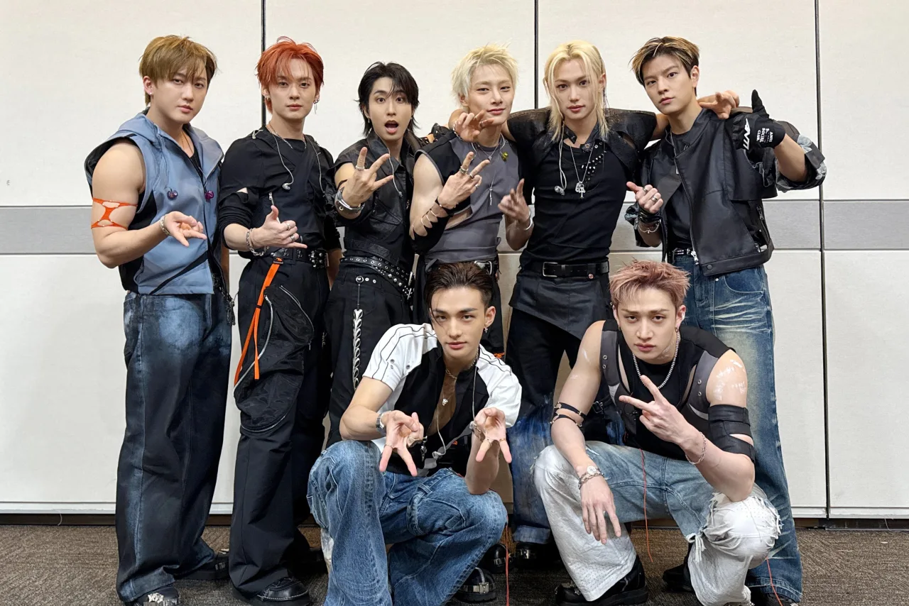 Membros do Stray Kids suando, segurando garrafas de água e fones, ao lado de um microfone no palco