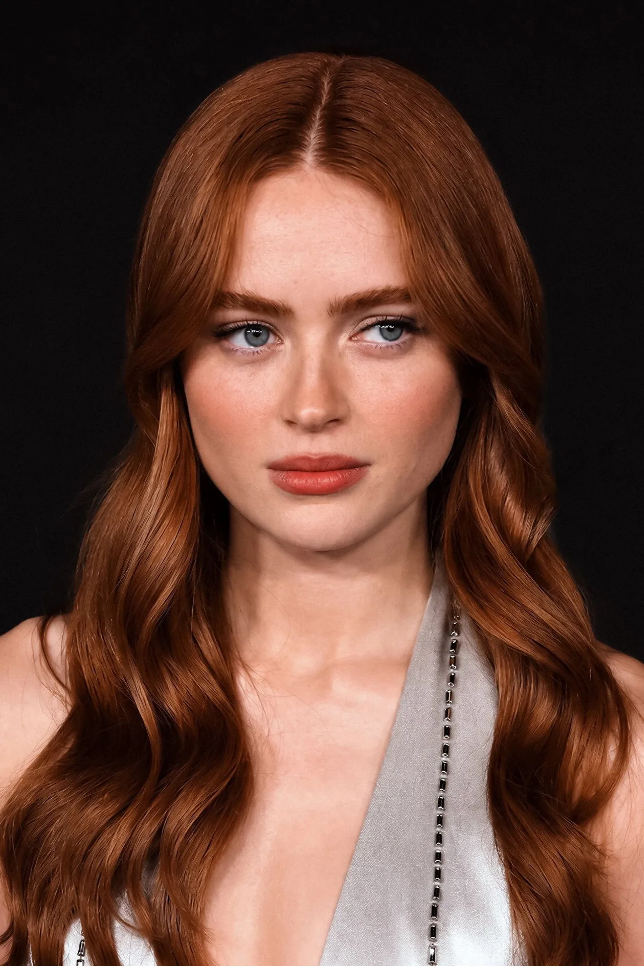 Sadie Sink vestindo roupa de treino, segurando halteres enquanto faz agachamento em uma academia iluminada