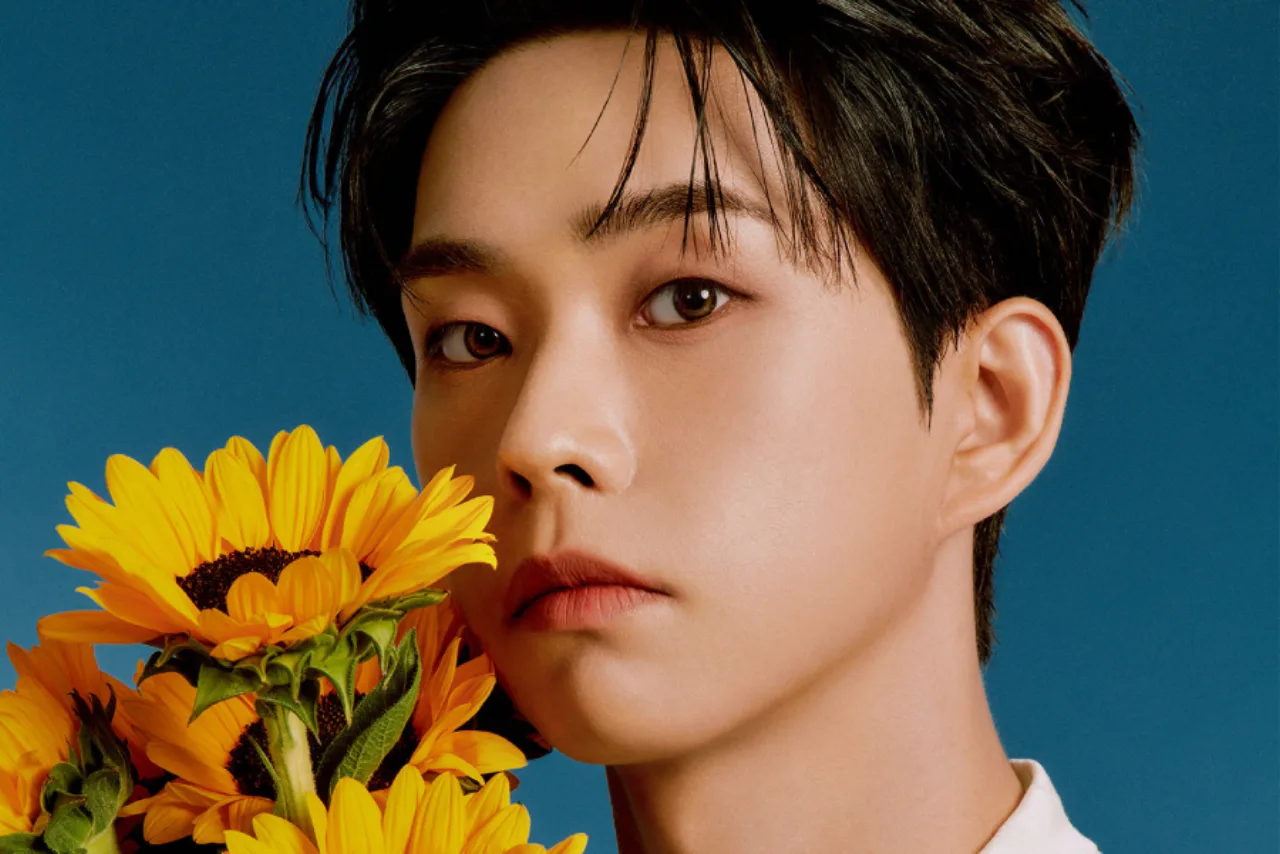 Choi Sang Yeop vestindo roupa de academia, segurando halteres, ao lado de um buquê de flores ao fundo