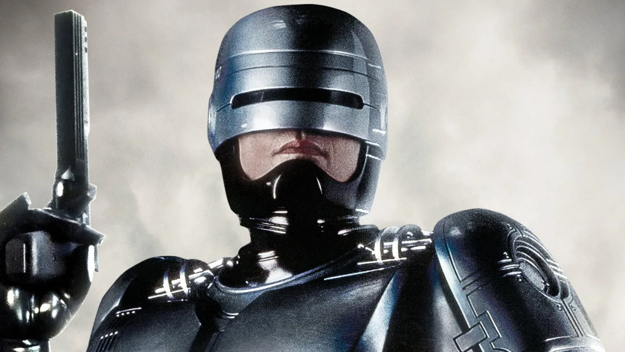Homem musculoso em camiseta do Robocop levanta halteres ao lado de garrafa de whey e tela exibindo a capa do filme