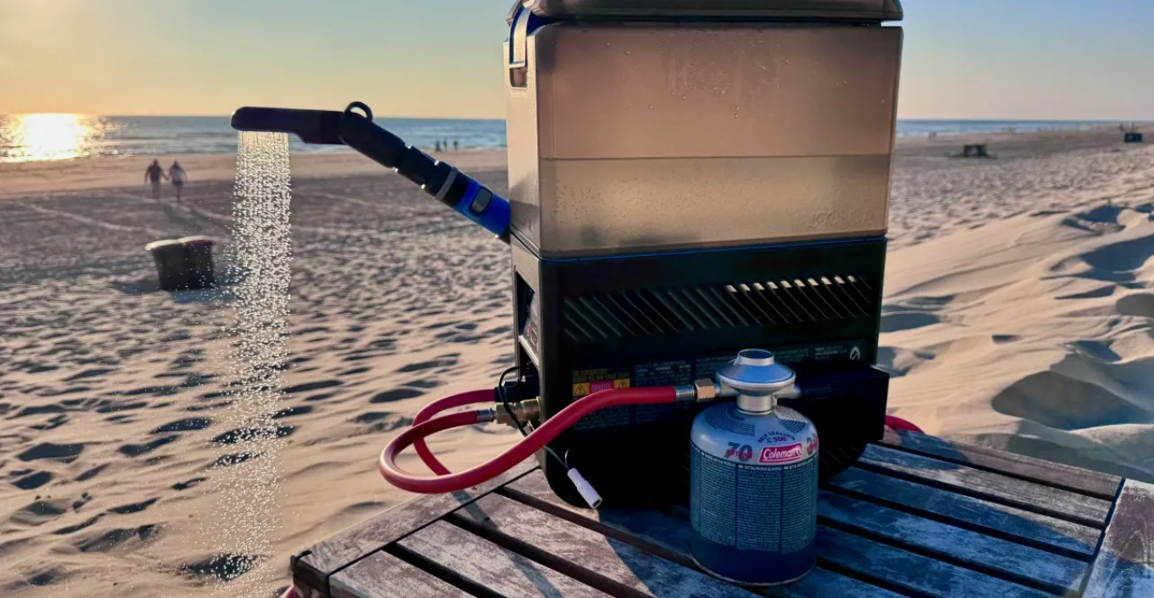 Homem usando ducha portátil ao ar livre, segurando garrafa de água e toalha, com mochila ao fundo