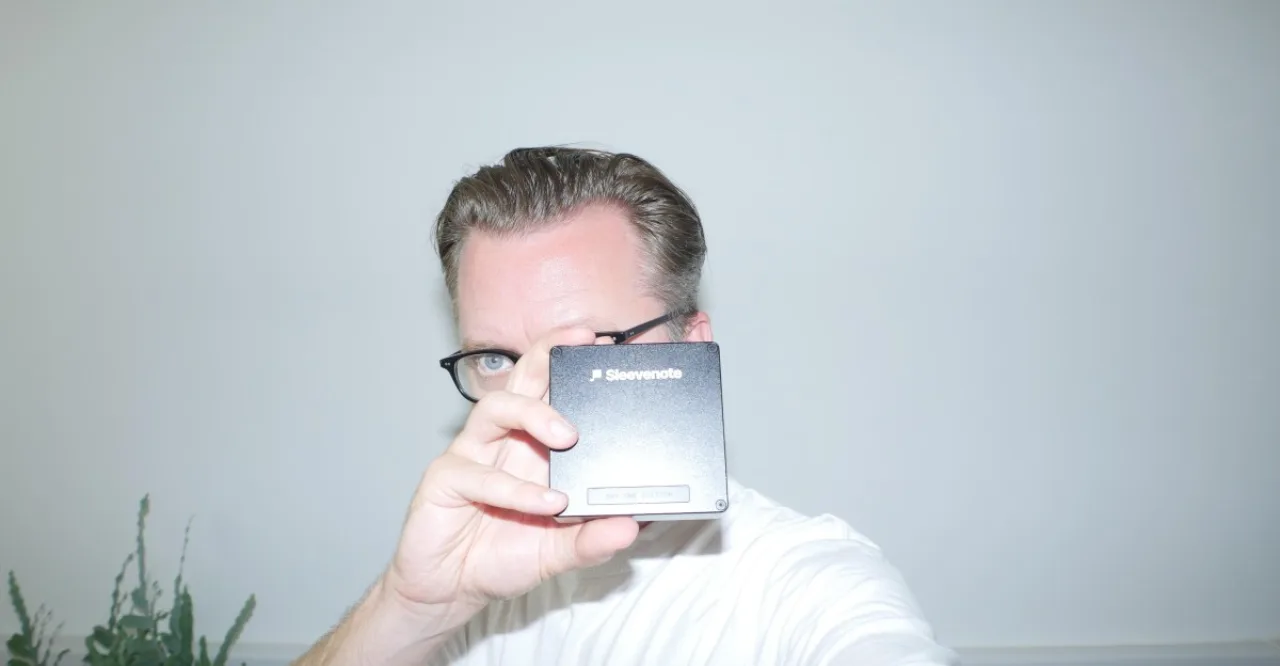 Tom Vek segurando um player digital ultrafino, com tela iluminada exibindo ondas sonoras e cabos de áudio ao fundo