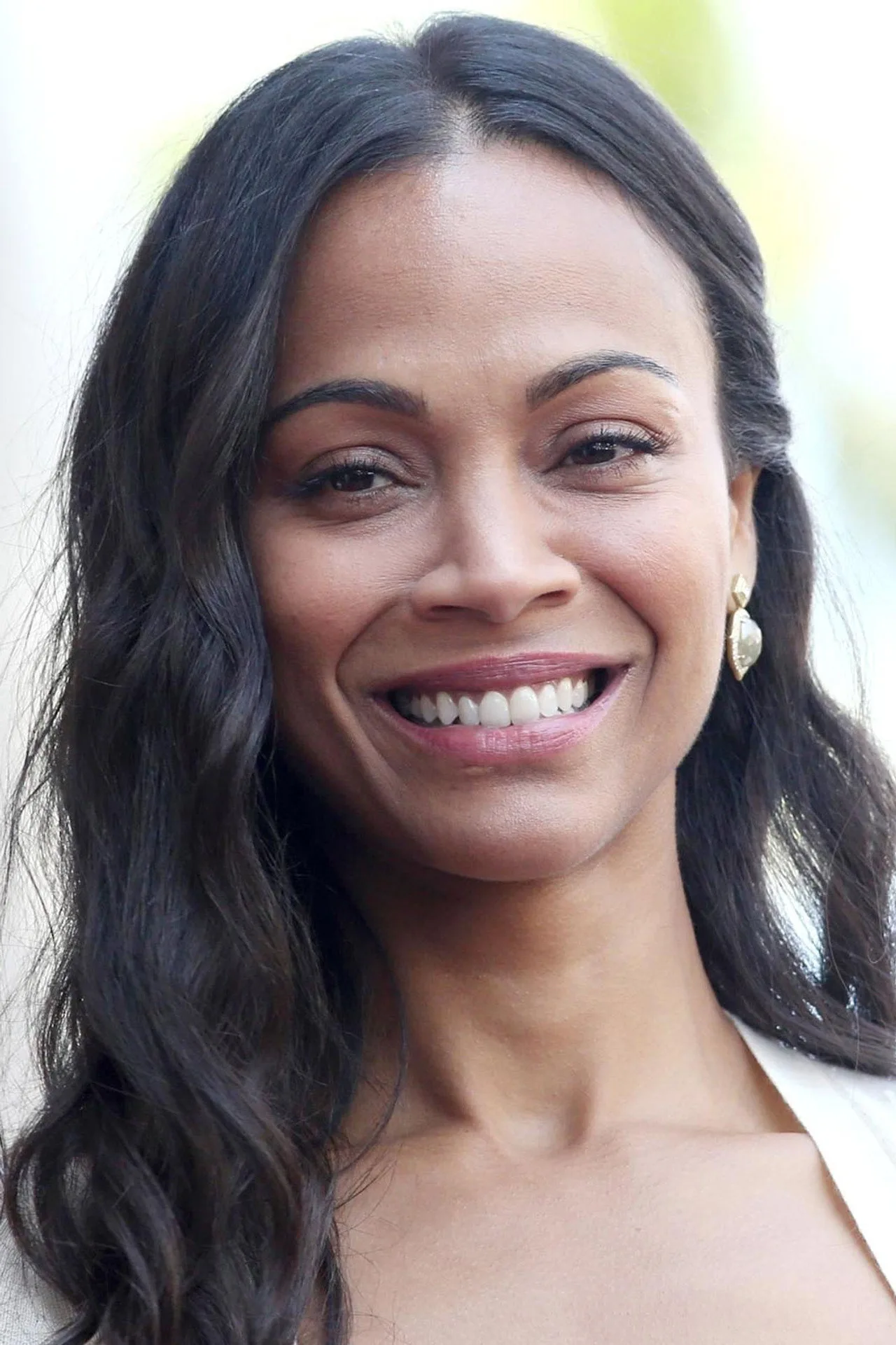Zoe Saldana executa agachamento com barra, vestindo legging cinza e top esportivo, ao lado de halteres e garrafa d'água