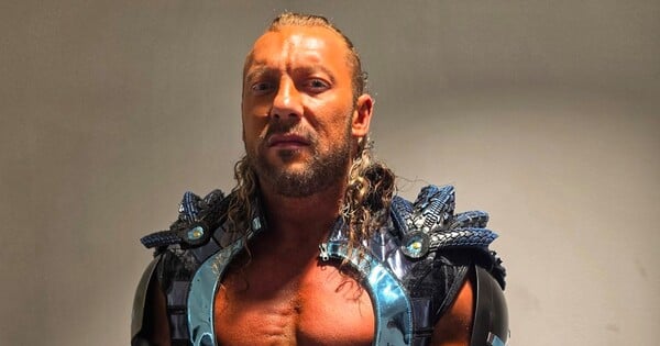 Kenny Omega em traje amarelo e vermelho de All Might, flexionando o bíceps com halteres na academia